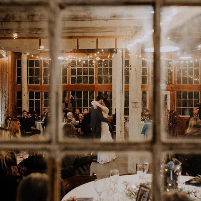 A bride and groom share their first dance, surrounded by wedding guests at a rustic indoor venue.