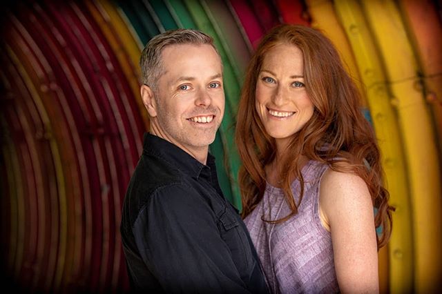 A smiling couple stands close together in front of a colorful, vibrant background in a studio setting.