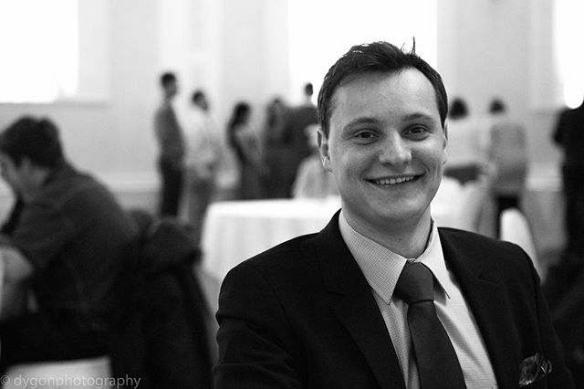 A smiling man in a suit attends an event. The black and white portrait captures a professional and confident atmosphere.
