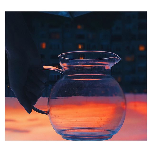 A glass pitcher reflects the orange sunset over a cityscape, held by a hand. A serene scene.