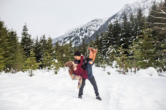 A couple shares a playful moment in a snowy winter landscape, surrounded by trees and mountains.