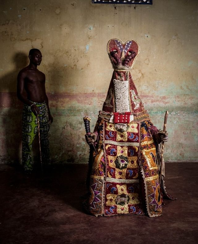 A man stands beside an elaborate masquerade costume embodying African cultural traditions.