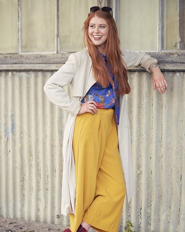 A happy redhead woman in yellow trousers and a beige coat stands outdoors.