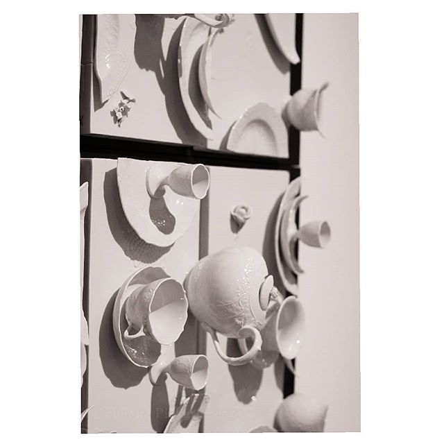 A monochromatic art installation featuring white ceramic cups, plates and teapots mounted on a wall, creating a sculptural display.