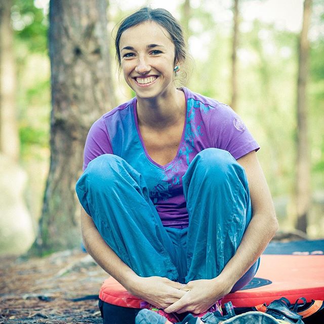 A smiling woman sitting cross-legged on a boulder mat in a forest.