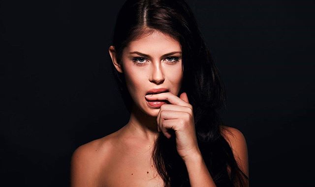 A brunette woman poses in a sensual close-up against a dark background.