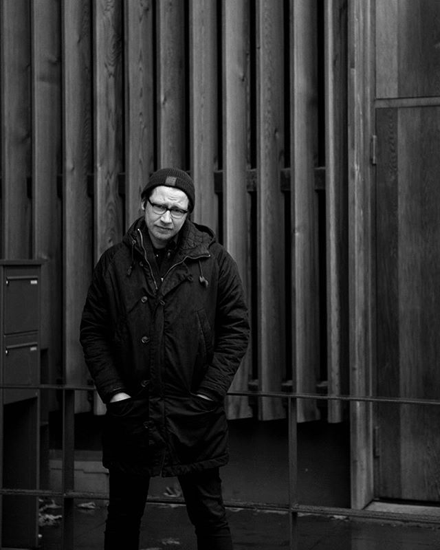 A man in a coat and beanie stands against a wooden backdrop in a black and white urban scene.