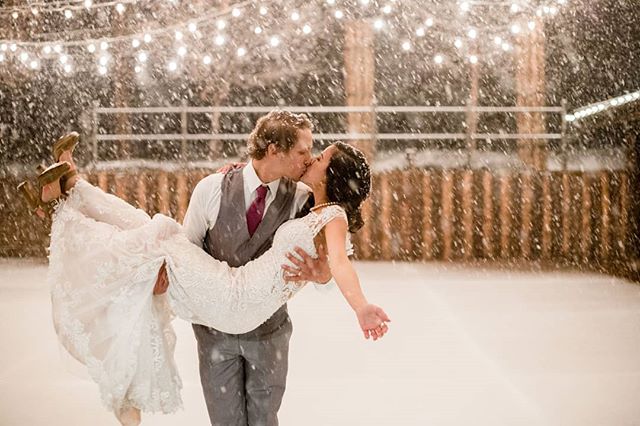 A bride and groom share a kiss during their winter wedding, surrounded by falling snow and warm, romantic lights.