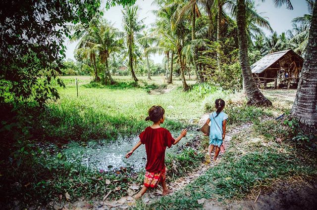 Two girls walk along a path through a lush, tropical rural landscape with palm trees and a small hut.
