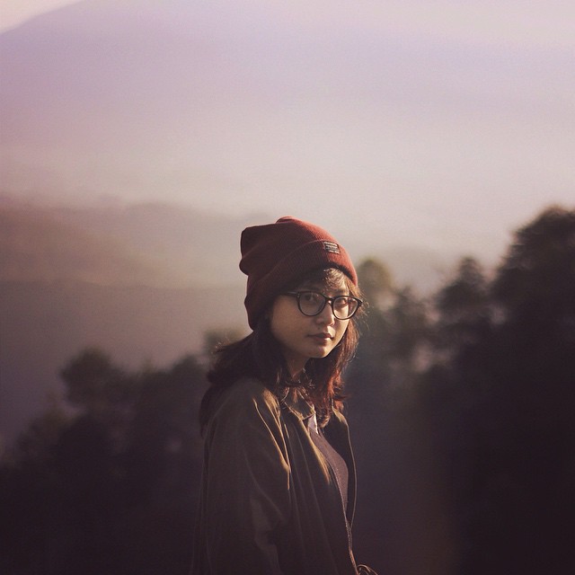 A woman wearing glasses and a beanie stands outdoors with mountains in the background.
