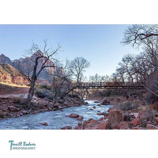A tranquil river scene with a bridge, mountains, and bare trees under a clear blue sky.