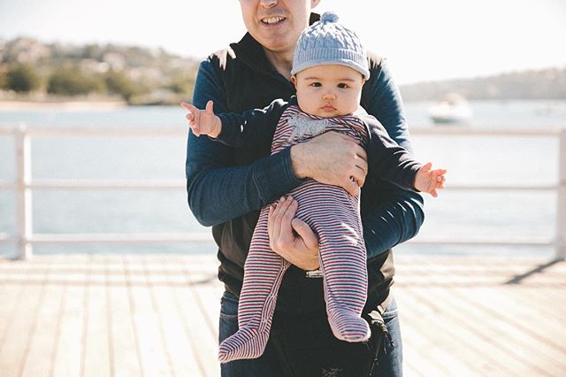 A father holds his baby on a pier, enjoying a sunny day with a beautiful water view.