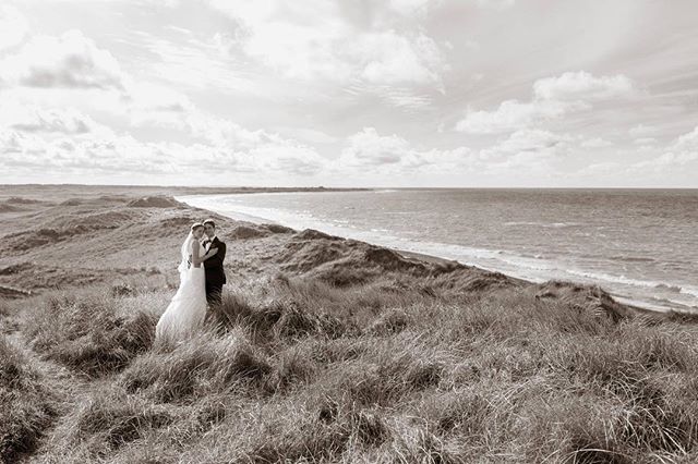 A bride and groom embrace on a grassy dune overlooking a beach and ocean in a vintage-style wedding photo.