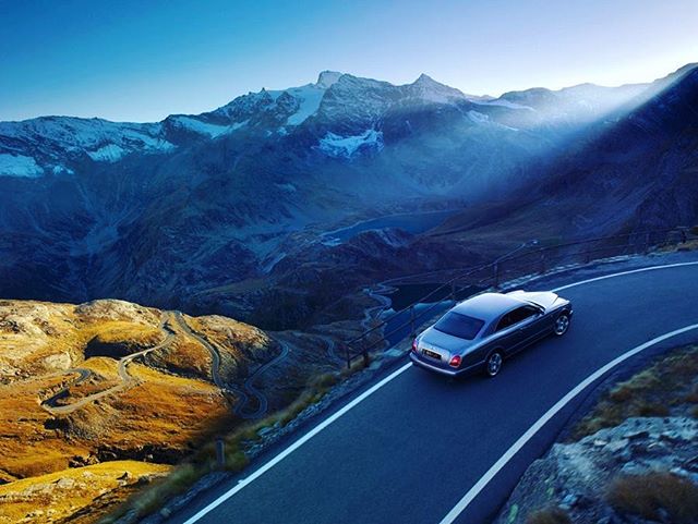 A car drives on a winding mountain road with stunning alpine scenery in the background, capturing a sense of adventure.