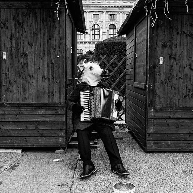 A street performer in a horse mask plays an accordion between two wooden stalls.