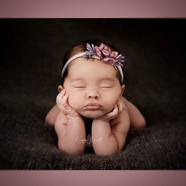 A peacefully sleeping newborn baby with a flower headband, resting on a soft brown blanket.