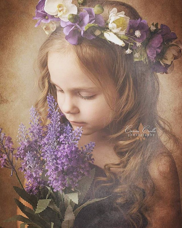 A young girl wearing a floral crown softly holds a bouquet of lavender in a dreamy, vintage-style portrait.