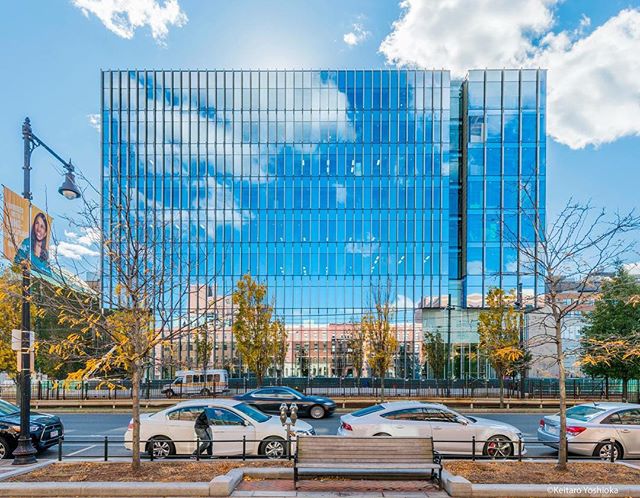 A modern glass building reflects the blue sky and clouds, cars drive by on the street below.