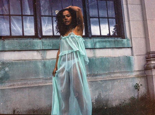 A woman in a sheer dress poses against an old building for a vintage fashion shoot.