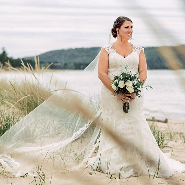 A bride in a lace wedding dress stands on a beach holding a bouquet, with a lake in the background.