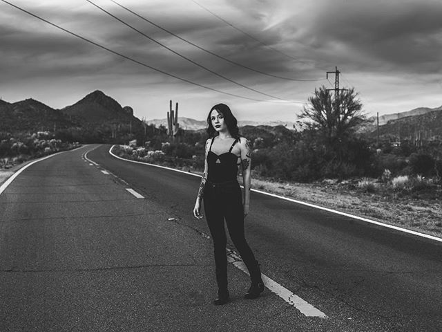 A woman stands on a desert road. Black and white image shows mountains in the background.