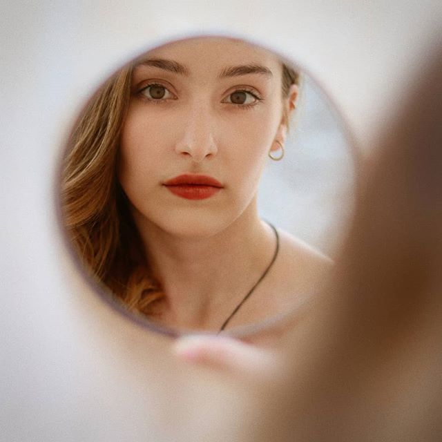 A woman is reflected in a round mirror, showcasing a close-up of her face with soft, natural lighting.