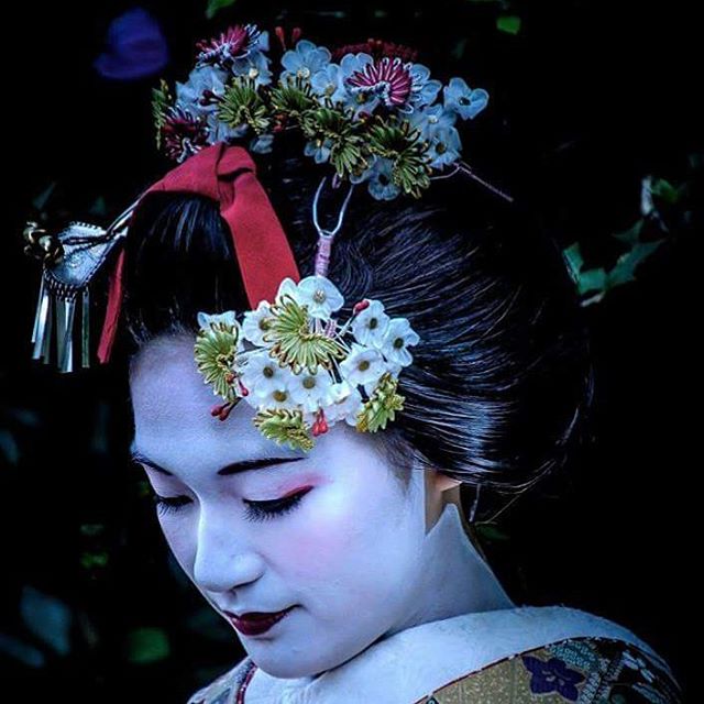 A geisha with pale makeup and elaborate floral headpiece is looking down in a dark, outdoor setting.