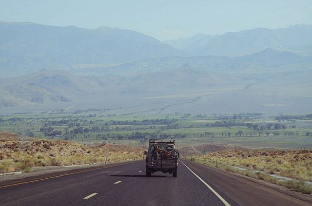 A car drives on a long highway with a bike mounted on the back, with mountains in the distance.