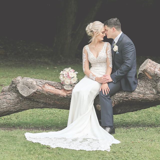 A bride and groom share a tender moment sitting on a log in a park on their wedding day.