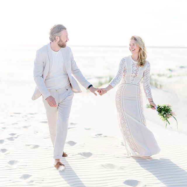 A happy couple walks hand-in-hand on a bright sandy beach on their wedding day, smiling at each other.