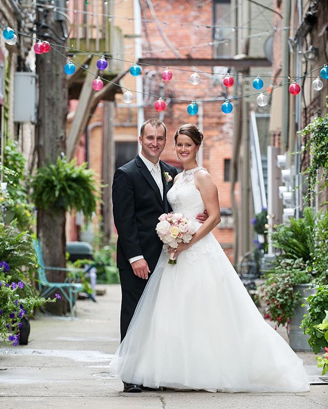 A bride and groom stand in an alley with string lights, holding a bouquet and smiling on their wedding day.