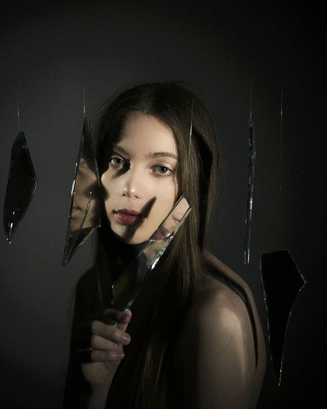 A woman poses with broken mirrors reflecting fragmented images in a moody studio portrait.