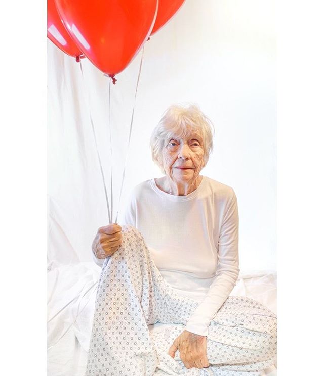 A happy senior woman sits holding red balloons in a bright, celebratory setting.
