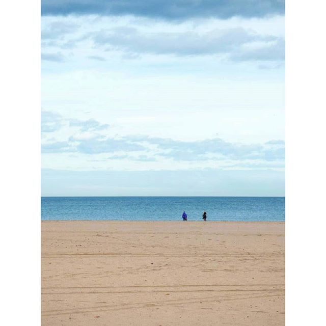 Two people stand on a sandy beach, facing the calm ocean under a cloudy sky.