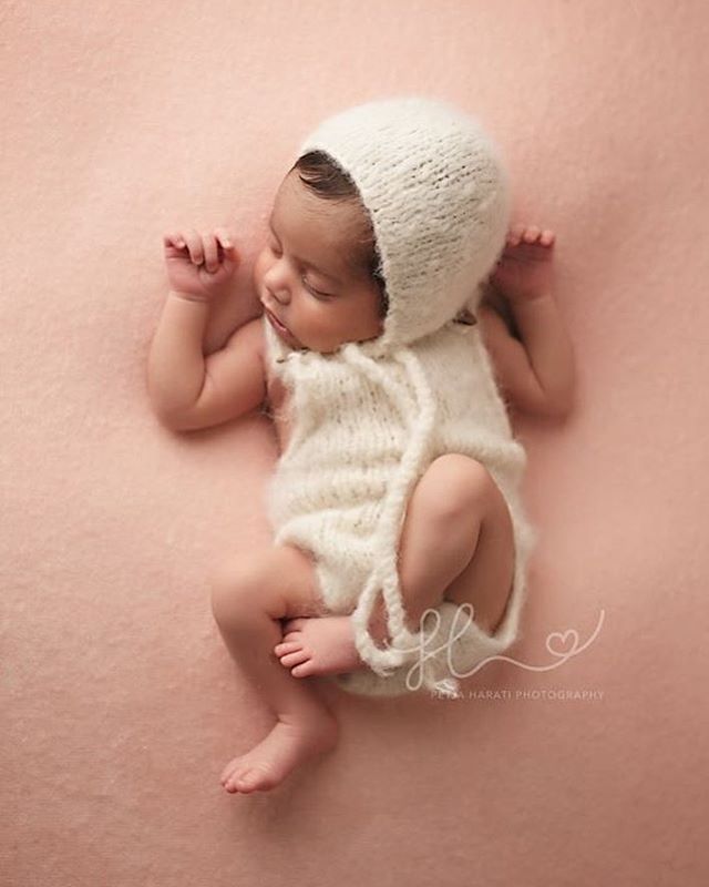 A newborn baby sleeps peacefully wrapped in soft, white knitwear against a peach-toned backdrop.