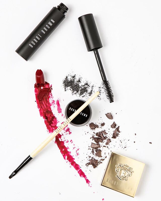 Flat lay of Bobbi Brown makeup products including lipstick, mascara, eye shadow, and brush on a white background.