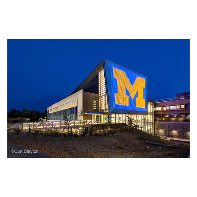 The University of Michigan building illuminated at night features a large "M" logo on the exterior.