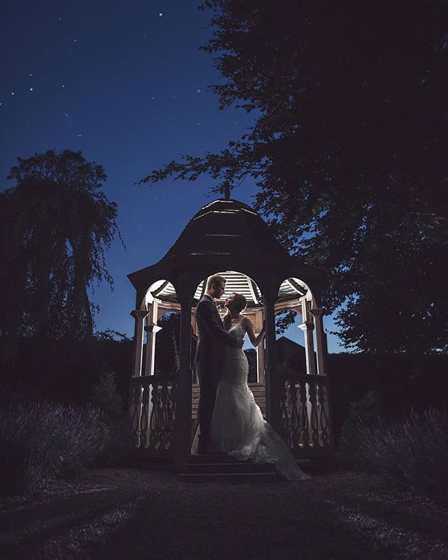 A bride and groom share a romantic moment in a gazebo at night, silhouetted against the evening sky.