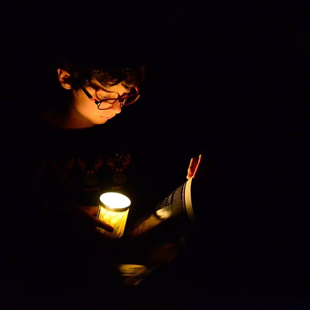 A boy with glasses reads a book by the light of a candle, creating a warm and intimate atmosphere in the dark.