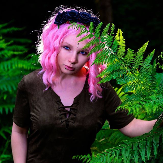 A woman with pink hair and a flower crown poses among ferns in a forest setting.
