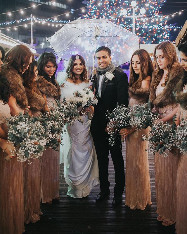 A bride, groom and their wedding party are smiling under an umbrella near a christmas tree at night.