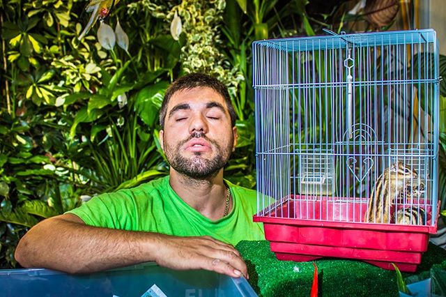 A man with closed eyes poses with a chipmunk in a cage, in front of a lush green background.
