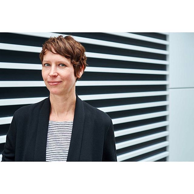 A woman with short brown hair smiles in front of a striped wall. She is wearing a black blazer and striped top.