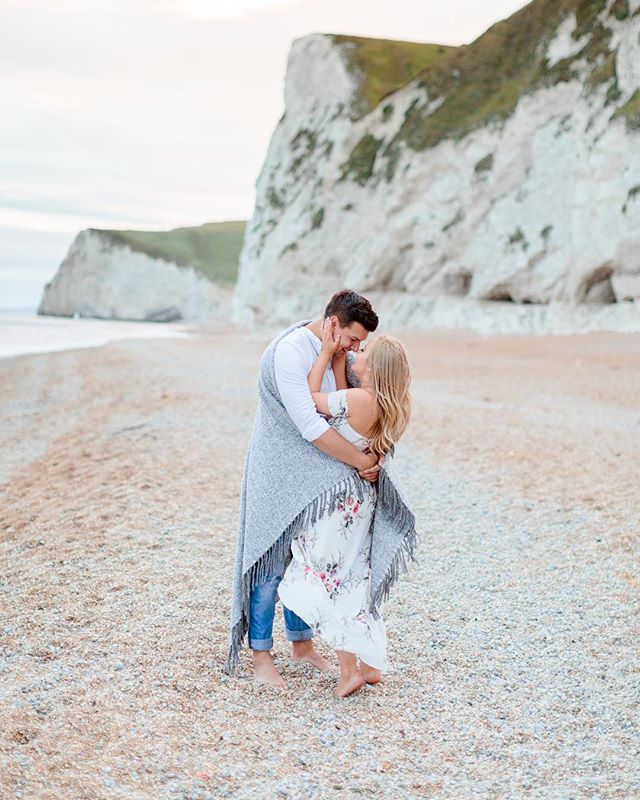A couple embraces on a pebble beach with cliffs in the background on a overcast day.
