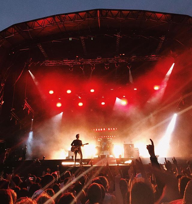 A musician plays guitar on stage at a concert, illuminated by red stage lights, with a large, enthusiastic crowd in attendance.