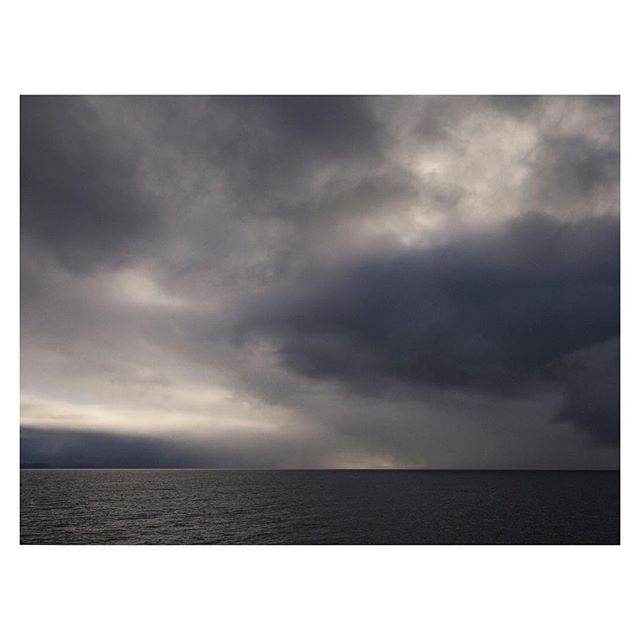 A dark, moody seascape featuring a calm sea under a dramatic, cloudy sky near the horizon.