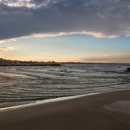 A tranquil beach scene with gentle waves and a cityscape under a cloudy sky.