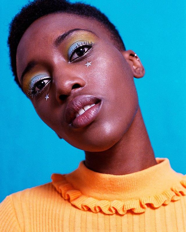 A vibrant portrait of a woman with creative makeup and star accents on a blue background.
