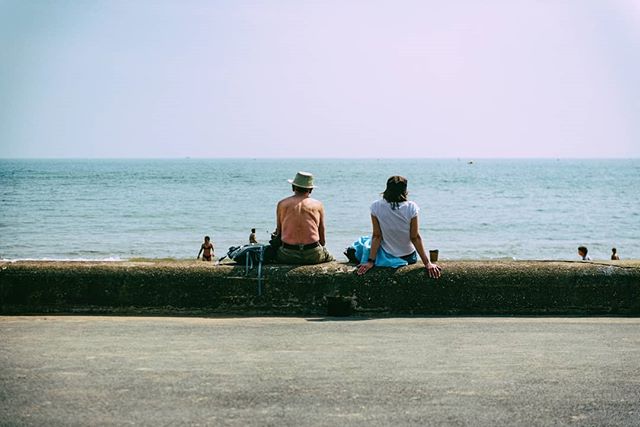 A couple sits on a seawall overlooking a calm ocean, enjoying a peaceful beach scene.