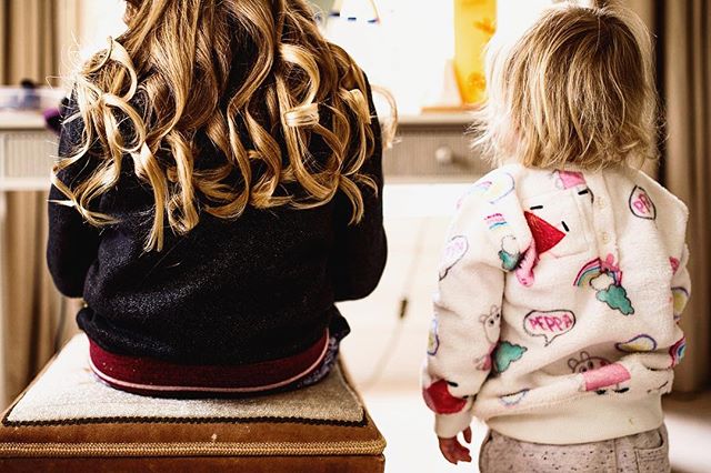 Two young girls are seen from the back, one with curly hair sitting on a stool.